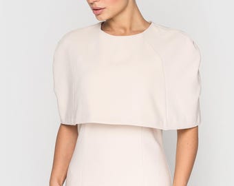 May include: A light beige dress with a cape-like overlay. The dress has a round neckline and a fitted silhouette. The cape sleeves extend to the upper arms. The model is wearing the dress against a white background.