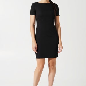 May include: A solid black short-sleeved dress featuring a boat neckline and a fitted silhouette that ends above the knee. The garment has clean, simple lines with subtle vertical seam detailing along the torso, providing a structured appearance.