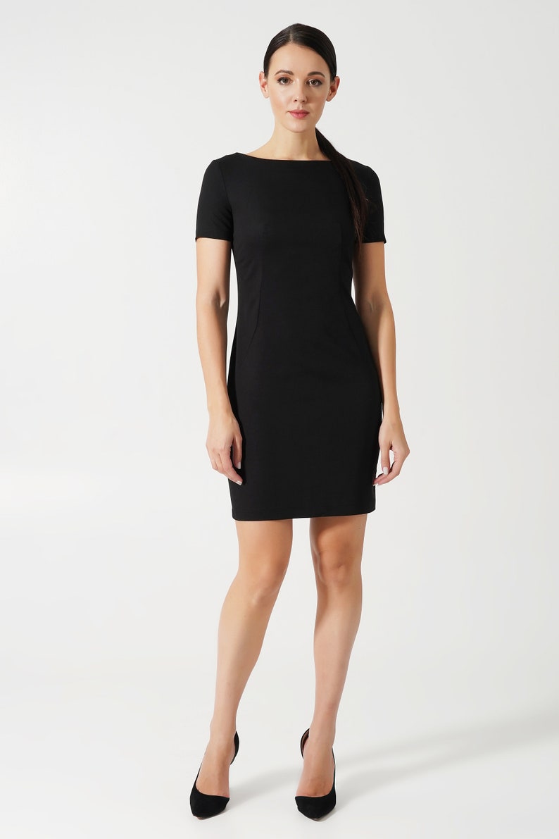 May include: A solid black short-sleeved dress featuring a boat neckline and a fitted silhouette that ends above the knee. The garment has clean, simple lines with subtle vertical seam detailing along the torso, providing a structured appearance.