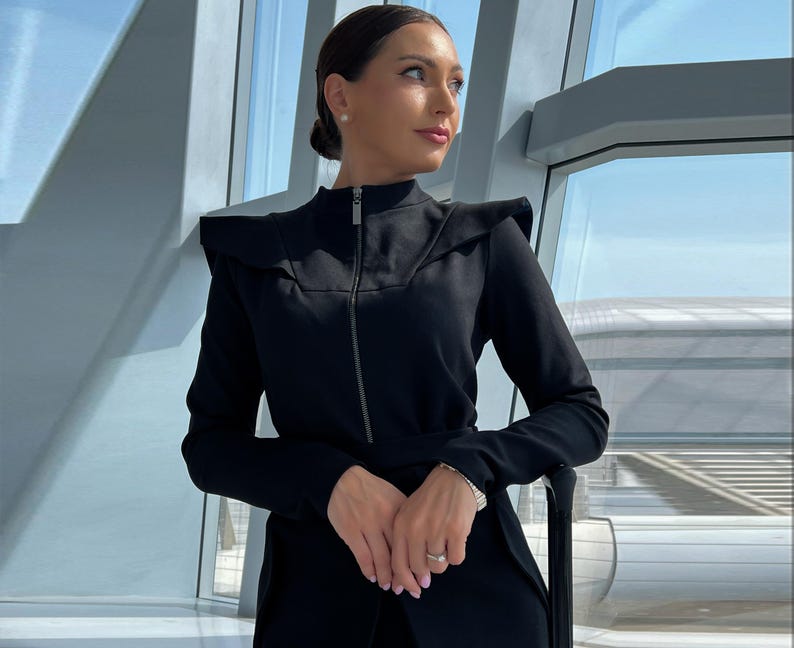 A black zip-up jacket with a high collar and a belt at the waist. The jacket has a ruffled detail on the shoulders.