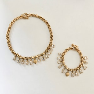 Puede incluir: Dos collares de oro con dijes. El collar más grande tiene la palabra "LOVE" escrita con dijes, y un dije en forma de corazón. El collar más pequeño tiene la palabra "SUMMER" escrita con dijes, y un dije en forma de corazón.