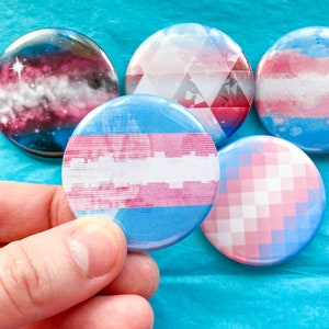 Op de afbeelding: Vijf ronde knopen met verschillende ontwerpen. De knopen zijn allemaal blauw, roze en wit. Eén knoop heeft een galaxy-ontwerp, één heeft een geruit ontwerp, één heeft een gestreept ontwerp en twee hebben geometrische ontwerpen.