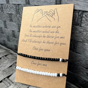 Puede incluir: Dos pulseras de amistad con cuentas, una negra y otra blanca, se muestran en una tarjeta marr&oacute;n. La tarjeta presenta un dibujo de manos haciendo una promesa de me&ntilde;ique y el texto "No matter where we go... One for you. One for me."