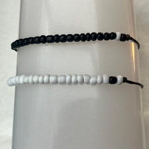 Puede incluir: Dos pulseras de cuentas. Una es negra con cuentas blancas en los extremos, la otra es blanca con cuentas negras en los extremos. Ambas est&aacute;n ensartadas en cordones negros.