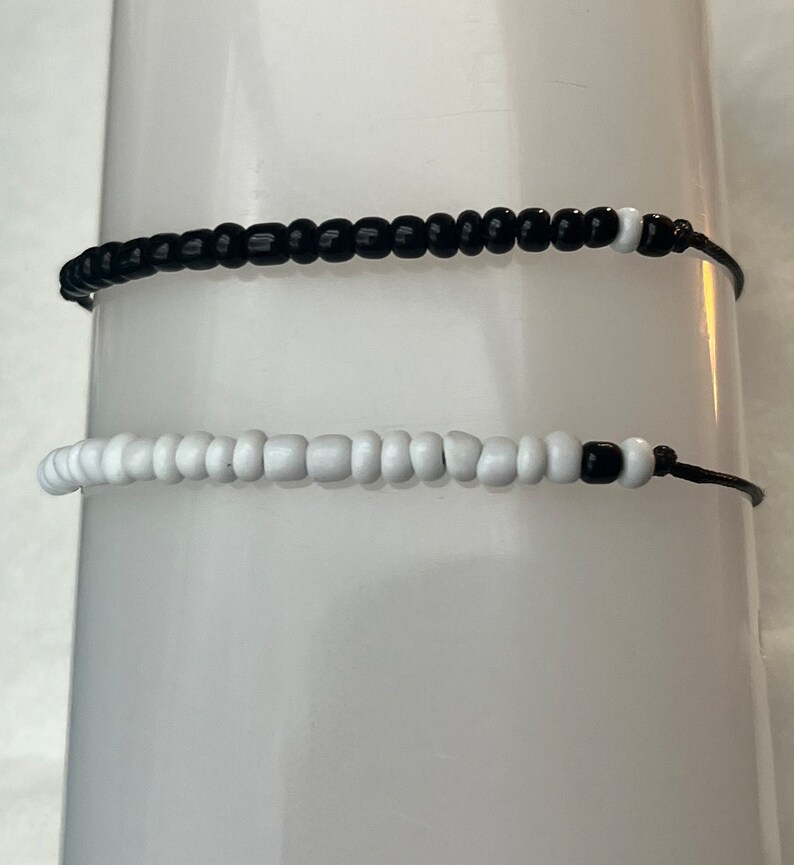Puede incluir: Dos pulseras de cuentas. Una es negra con cuentas blancas en los extremos, la otra es blanca con cuentas negras en los extremos. Ambas est&aacute;n ensartadas en cordones negros.