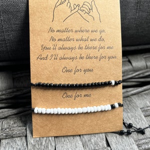 Puede incluir: Un par de pulseras de amistad, una negra y otra blanca, expuestas en una tarjeta marr&oacute;n. La tarjeta presenta un dibujo de dos manos haciendo una promesa de me&ntilde;ique y el texto "No matter where we go... One for you, One for me."