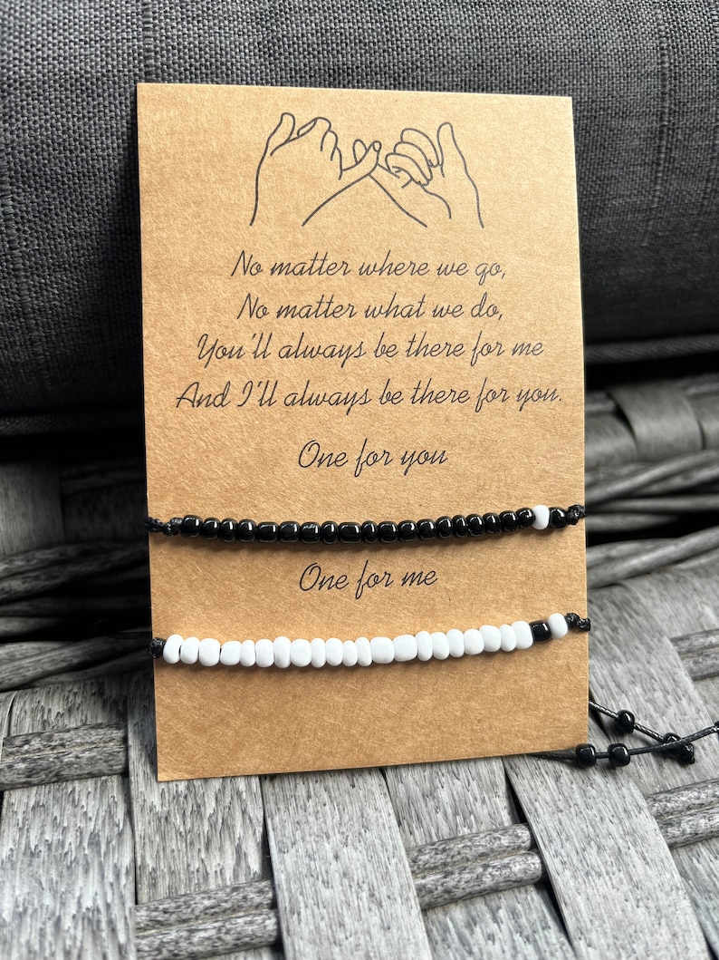 Puede incluir: Un par de pulseras de amistad, una negra y otra blanca, expuestas en una tarjeta marr&oacute;n. La tarjeta presenta un dibujo de dos manos haciendo una promesa de me&ntilde;ique y el texto "No matter where we go... One for you, One for me."