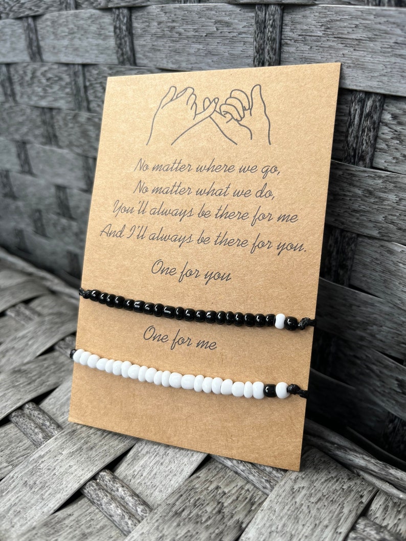 Puede incluir: Dos pulseras de amistad con cuentas, una negra y otra blanca, se muestran en una tarjeta marr&oacute;n. La tarjeta presenta un dibujo de manos haciendo una promesa de me&ntilde;ique y el texto "No matter where we go... One for you. One for me."