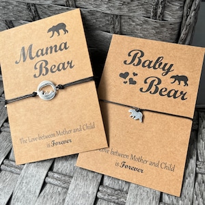 Può includere: Due etichette di cartoncino marrone con un cordoncino nero attaccato. L'etichetta superiore recita "Mama Bear" con una piccola sagoma di orso e un ciondolo con due orsi in un cerchio. L'etichetta inferiore recita "Baby Bear" con tre piccoli cuori e un ciondolo con un orso. Entrambe le etichette hanno il testo "L'amore tra madre e figlio è per sempre".