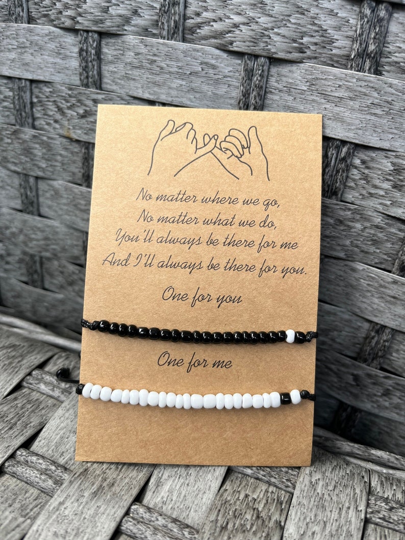 Puede incluir: Dos pulseras de amistad con cuentas, una negra y otra blanca, se muestran en una tarjeta marr&oacute;n. La tarjeta presenta un dibujo lineal de dos manos haciendo una promesa me&ntilde;ique y el texto: "No importa a d&oacute;nde vayamos... Uno para ti, Uno para m&iacute;."
