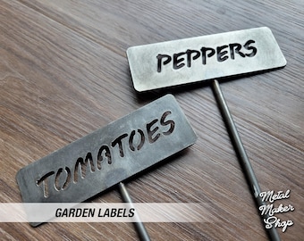 May include: Two metal garden labels with black lettering. The labels are attached to metal stakes. The top label reads "PEPPERS" and the bottom label reads "TOMATOES". The labels are on a wooden surface.  GARDEN LABELS is printed below the labels.