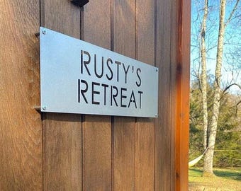 May include: A silver metal sign with the words "Rusty's Retreat" cut out in black lettering. The sign is attached to a wooden door.