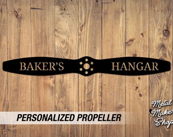 May include: Black metal propeller with "BAKER'S HANGAR" engraved in gold lettering. The propeller is mounted on a wood background.  Personalized Propeller.