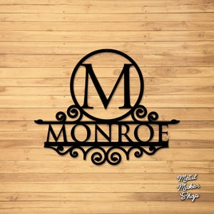 May include: Black metal wall art featuring the letter "M" inside a circle, above the name "MONROE". The design includes decorative scrollwork and is mounted on a wooden background. The text "Metal Maker Shop" is in the lower right corner.