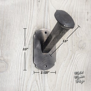 May include: A black metal wall hook shaped like a nail with a rectangular base. The hook measures 3.5 inches in height and 2.125 inches in width. The hook is mounted on a white wooden surface.