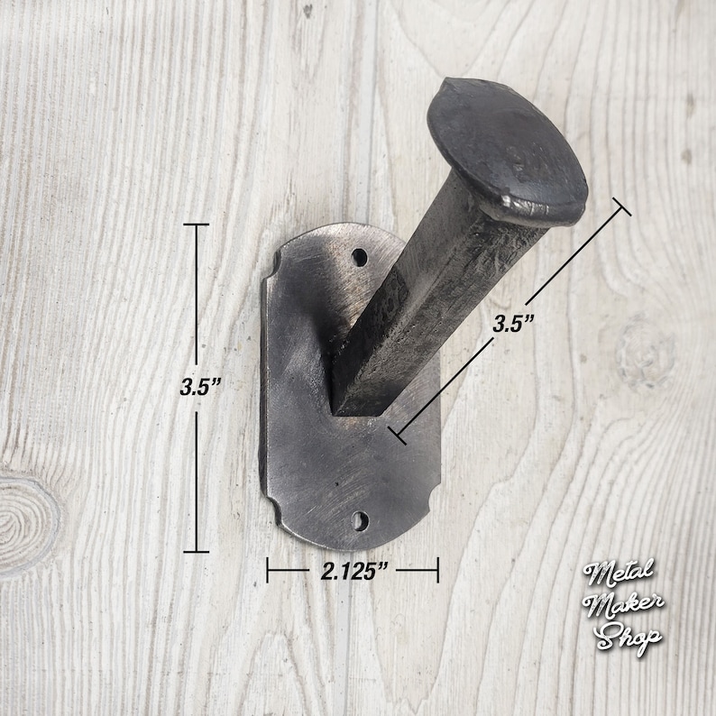 May include: A black metal wall hook shaped like a nail with a rectangular base. The hook measures 3.5 inches in height and 2.125 inches in width. The hook is mounted on a white wooden surface.