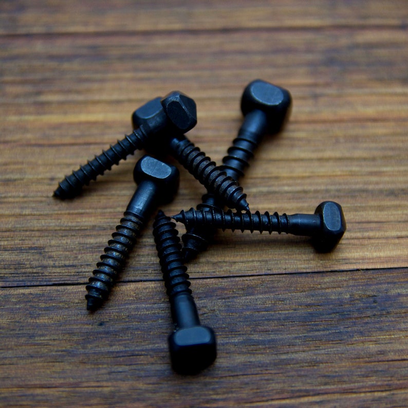 May include: A group of six black metal screws with square heads. The screws are arranged in a star shape on a wooden surface.