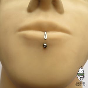 Può includere: Un gioiello per piercing labret verticale color argento con un design a barra curva. L'estremità superiore è un pezzo liscio e allungato a forma di goccia, mentre l'estremità inferiore è una piccola sfera metallica rotonda con una gemma chiara.