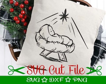 May include: A beige pillow with a black line drawing of baby Jesus in a manger, beneath a shining star. The pillow is in a wooden basket with red berries and greenery. Text on the bottom reads "SVG Cut File".