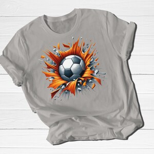 May include: A light gray t-shirt featuring a soccer ball design. The soccer ball is white and black, bursting through an orange and gray graphic. The shirt is made of a soft material.