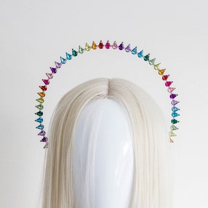 May include: A rainbow-colored halo crown with teardrop-shaped gems. The gems are arranged in a semi-circle, with colors transitioning from red to purple. The crown is placed on a mannequin head with long, blonde hair.