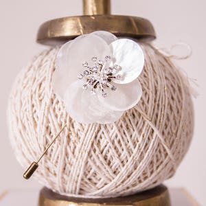 May include: A white pearl flower brooch with a rhinestone center, pinned to a ball of white yarn on a gold stand.