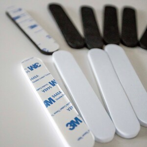 May include: An assortment of adhesive strips, some white and some black, are arranged on a white surface. Several white strips have blue and white 3M branding. The strips are designed for double-coated tissue tape.