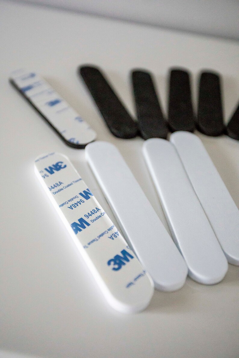 May include: An assortment of adhesive strips, some white and some black, are arranged on a white surface. Several white strips have blue and white 3M branding. The strips are designed for double-coated tissue tape.