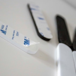 May include: Close-up of white adhesive strips with the blue "3M" logo and text. The strips are double-sided and designed for mounting. The image shows the adhesive backing being peeled away, revealing the sticky surface. Several strips are visible.
