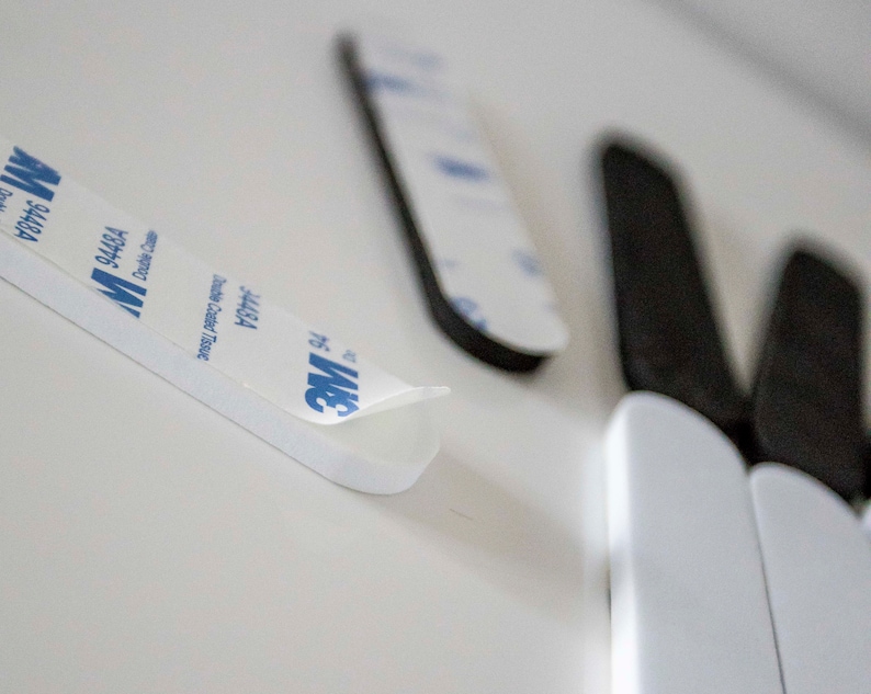 May include: Close-up of white adhesive strips with the blue "3M" logo and text. The strips are double-sided and designed for mounting. The image shows the adhesive backing being peeled away, revealing the sticky surface. Several strips are visible.