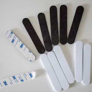 May include: An assortment of black and white adhesive strips, along with two strips of double-sided tape. The strips are oval-shaped and appear to be made of a smooth material. The tape has blue and white branding.