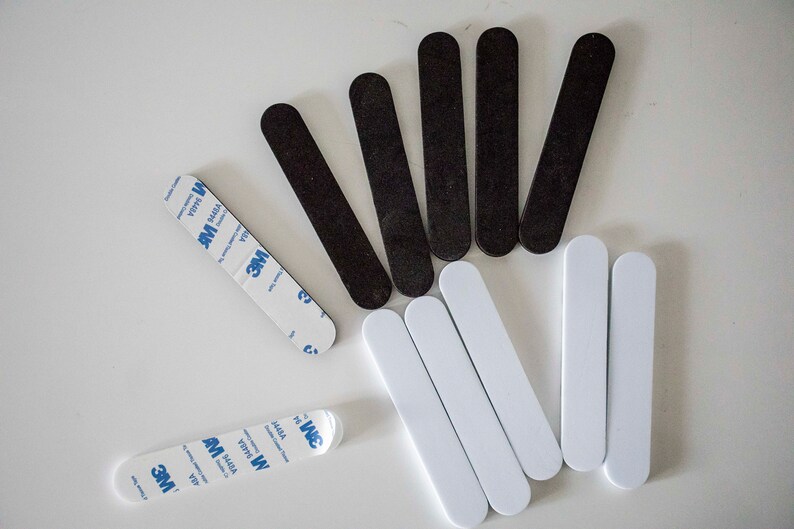 May include: An assortment of black and white adhesive strips, along with two strips of double-sided tape. The strips are oval-shaped and appear to be made of a smooth material. The tape has blue and white branding.