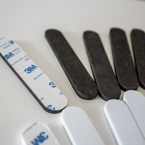 May include: An assortment of adhesive strips in black and white, with a 3M brand label visible on some. The strips are rectangular with rounded ends, designed for mounting or securing items. The image is shot from a high angle.