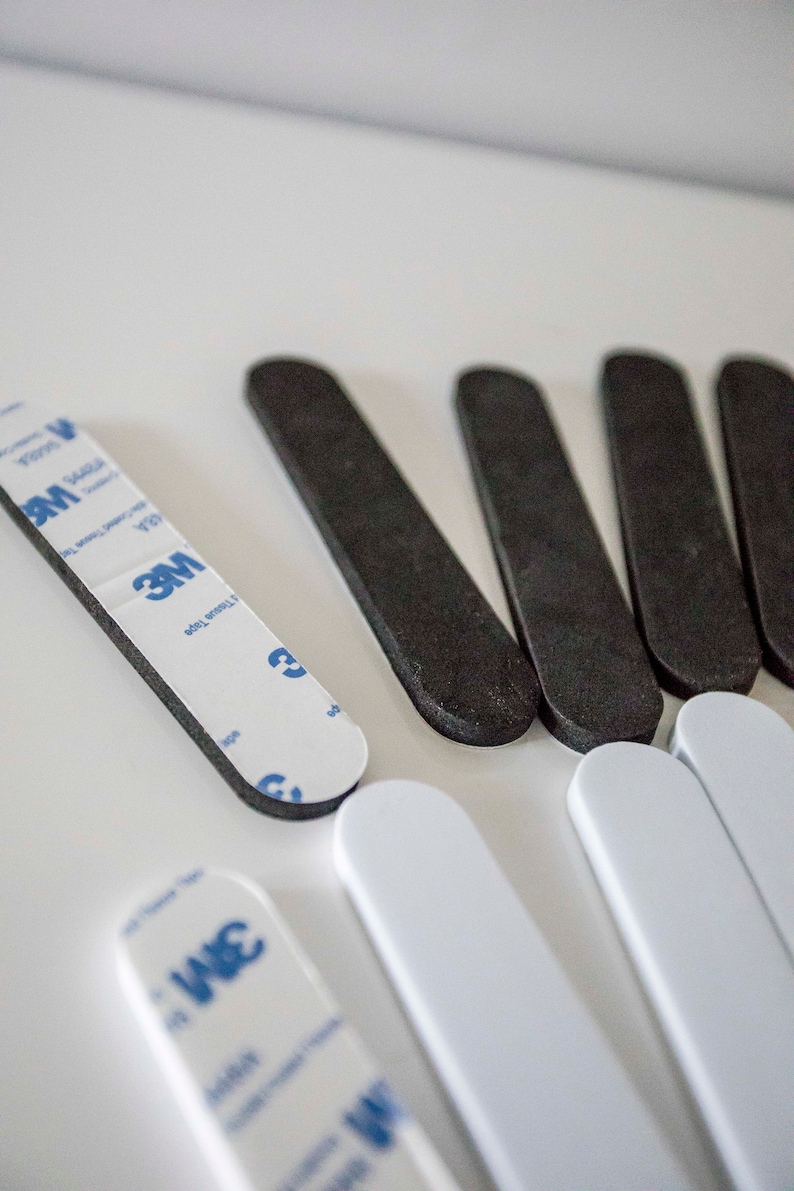 May include: An assortment of adhesive strips in black and white, with a 3M brand label visible on some. The strips are rectangular with rounded ends, designed for mounting or securing items. The image is shot from a high angle.