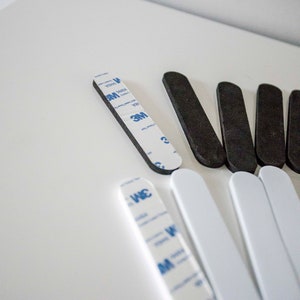 May include: An assortment of black and white adhesive strips, some with 3M branding. The strips are rectangular with rounded ends, designed for mounting or securing items. The image is taken from a high angle.