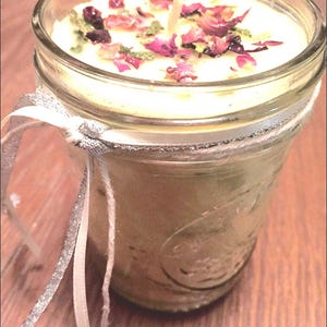 May include: A clear glass jar candle with a white wax base and a single wick. The candle is decorated with dried rose petals and green herbs.