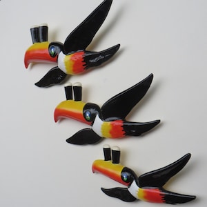 May include: Three toucan wall decorations with black wings, yellow beaks, red and white markings, and two black beer bottle tops on each beak.