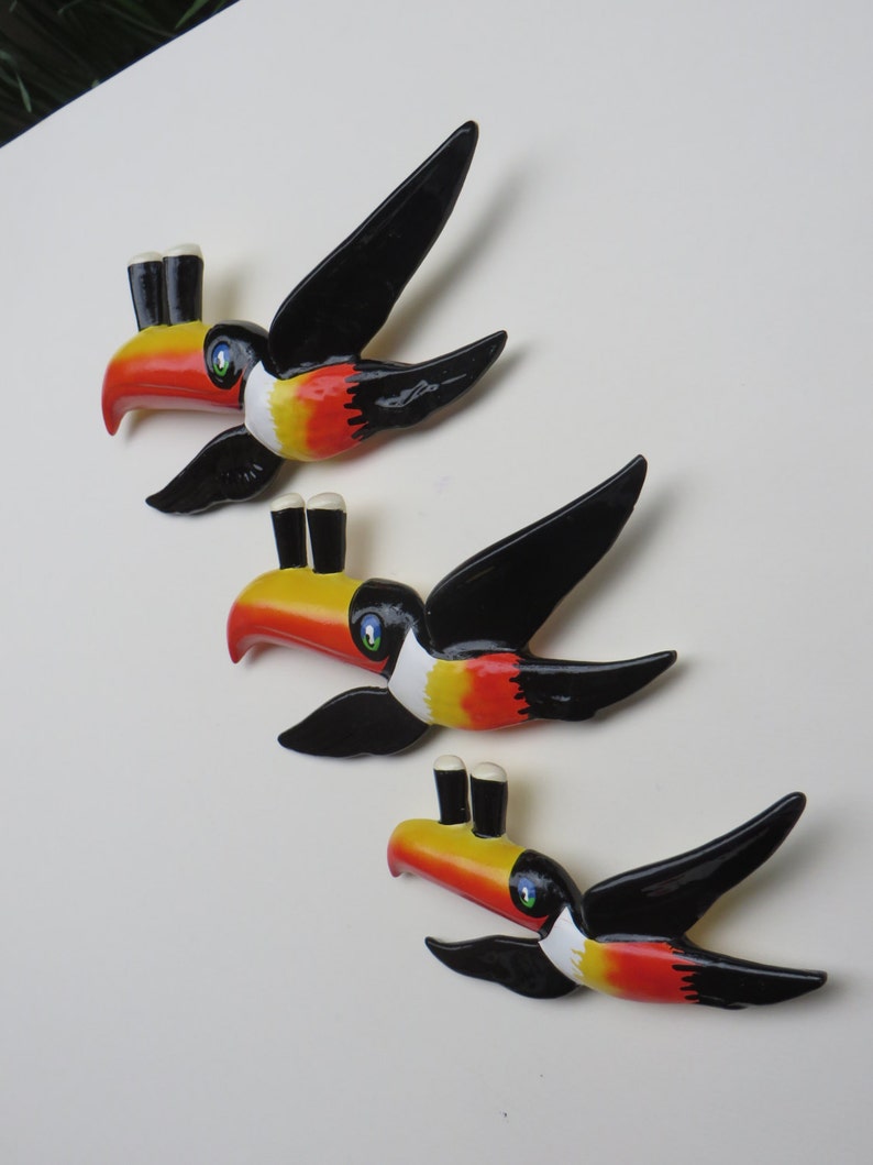 May include: Three toucan wall decorations with black wings, yellow beaks, red and white markings, and two black beer bottle tops on each beak.