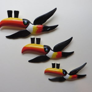 May include: Three toucan wall decorations with black wings, orange beaks, and white and yellow accents. Each toucan has two black pint glasses on top of its beak.