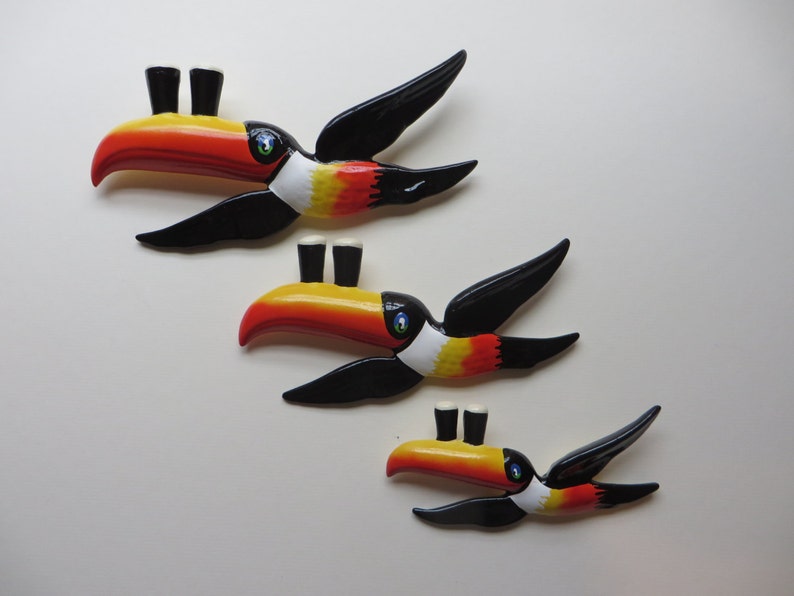 May include: Three toucan wall decorations with black wings, orange beaks, and white and yellow accents. Each toucan has two black pint glasses on top of its beak.