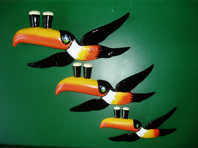 May include: Three toucan bird wall decorations with black wings, orange beaks, and white stripes. Each toucan is holding a pint of beer.
