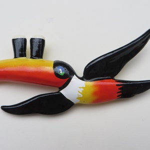 May include: A toucan figurine with a black body, red and yellow beak, and white chest. The toucan has two small black and white pint glasses for eyes.