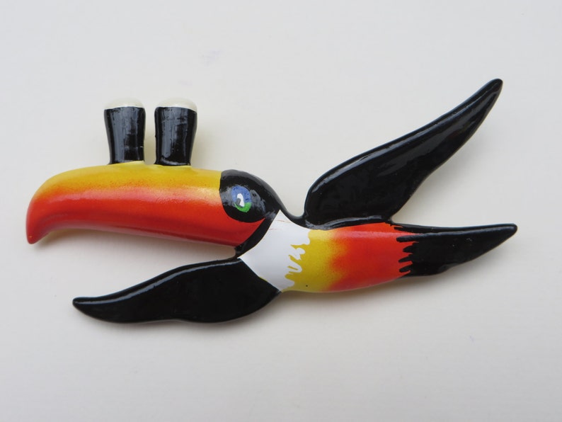 May include: A toucan figurine with a black body, red and yellow beak, and white chest. The toucan has two small black and white pint glasses for eyes.