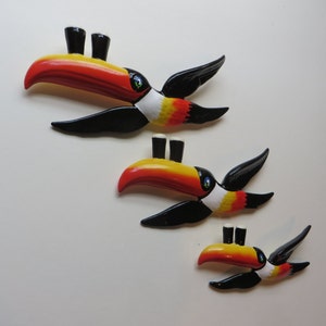May include: Three toucan-shaped wall decorations, each with a different size. The toucans are painted in black, white, yellow, and orange. Each toucan has two small black and white pint glasses on its beak.