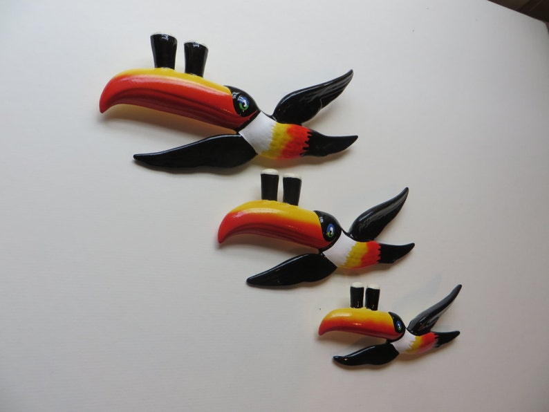 May include: Three toucan-shaped wall decorations, each with a different size. The toucans are painted in black, white, yellow, and orange. Each toucan has two small black and white pint glasses on its beak.