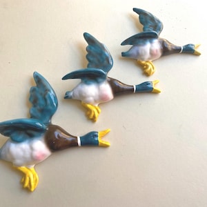 Könnte beinhalten: Drei fliegende Keramik-Entenfiguren. Die Enten haben blaue Flügel, braune Körper, weiße Brust, rosa Akzente und gelbe Füße. Jede Ente hat einen offenen Schnabel und ein weißes Halsband. Diese dekorativen Figuren sind wahrscheinlich für die Inneneinrichtung.
