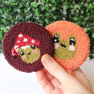 Pode incluir: Dois ornamentos de cogumelo redondos de feltro, costurados à mão. Um é um cogumelo bordô com um chapéu vermelho e um rosto verde. O outro é um cogumelo cor de pêssego com um rosto verde.