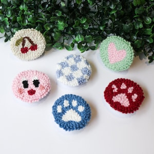 May include: Six colourful phone grips with different designs. The designs include a cherry, a heart, a gingham pattern, a cartoon face, and two paw prints.