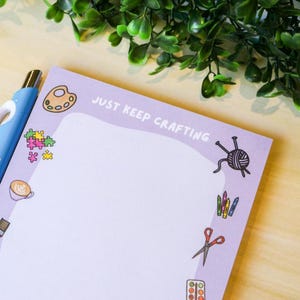 May include: A lavender notepad with the text "JUST KEEP CRAFTING" and craft-themed illustrations. A blue pen rests on the notepad. The notepad features illustrations of a paint palette, yarn, scissors, and other craft supplies.