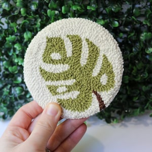 Pode incluir: Um descanso de copo redondo, branco, de feltro de agulha, com um design de folha de monstera verde.